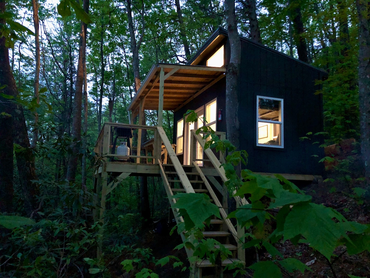 The treehouse is perched among trees, built on stilts with steps leading up to a covered porch. Large windows illuminate the interiors, while a wooden deck offers space for seating amidst the surrounding foliage.