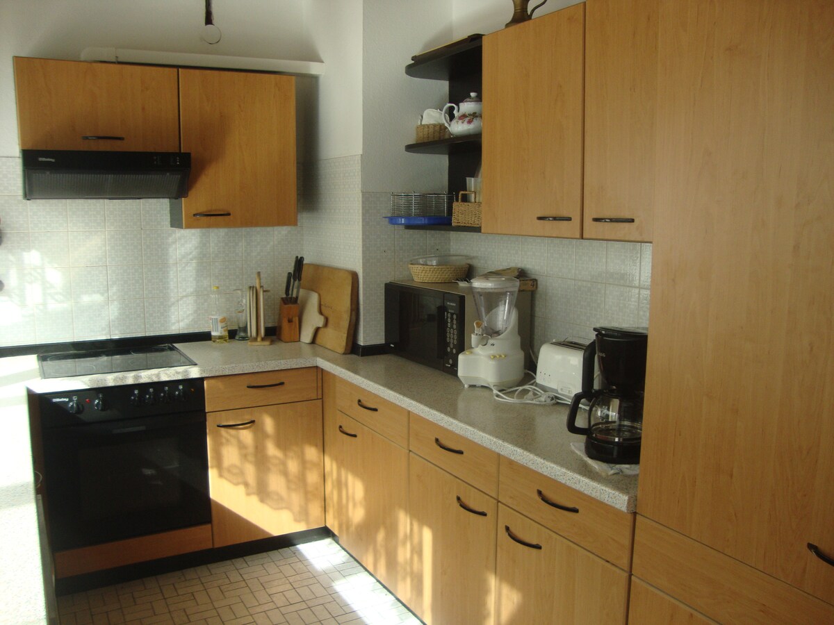 A modern kitchen is showcased with wooden cabinets and a light countertop. Essential appliances, including an oven, microwave, coffee maker, and blender, are visible. Natural light filters through a nearby window, illuminating the well-organized space.
