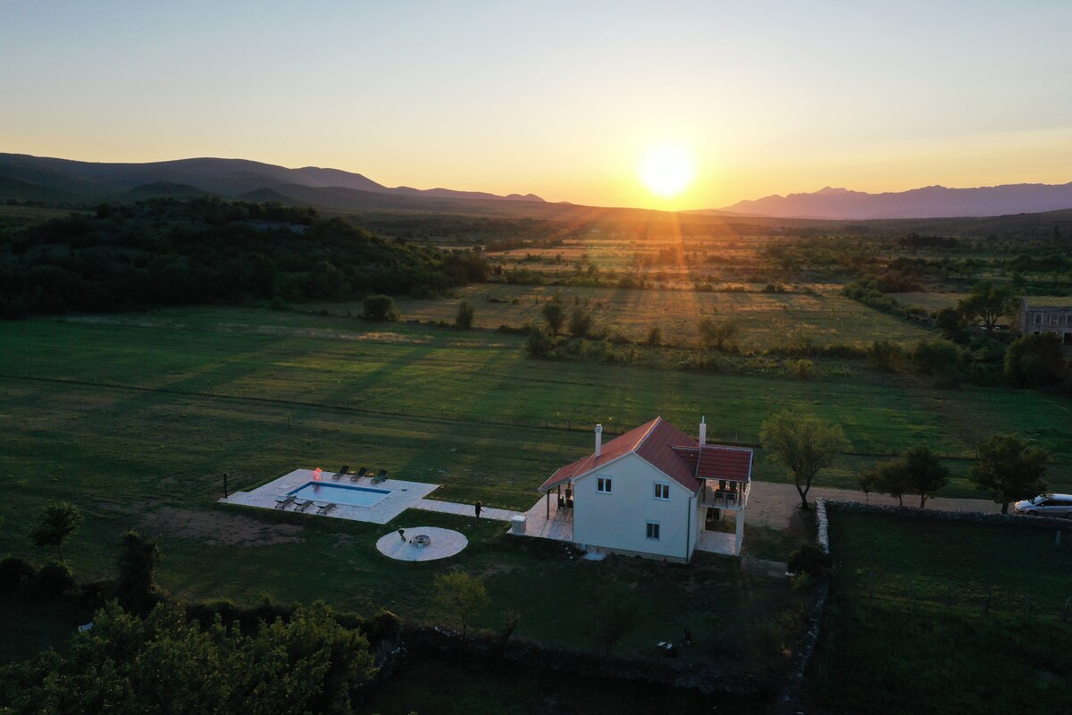 An aerial view captures a country house nestled in a vast green landscape at sunset. The structure features a red-tiled roof and is accompanied by a private swimming pool, while the horizon showcases distant mountains under a vibrant sky.