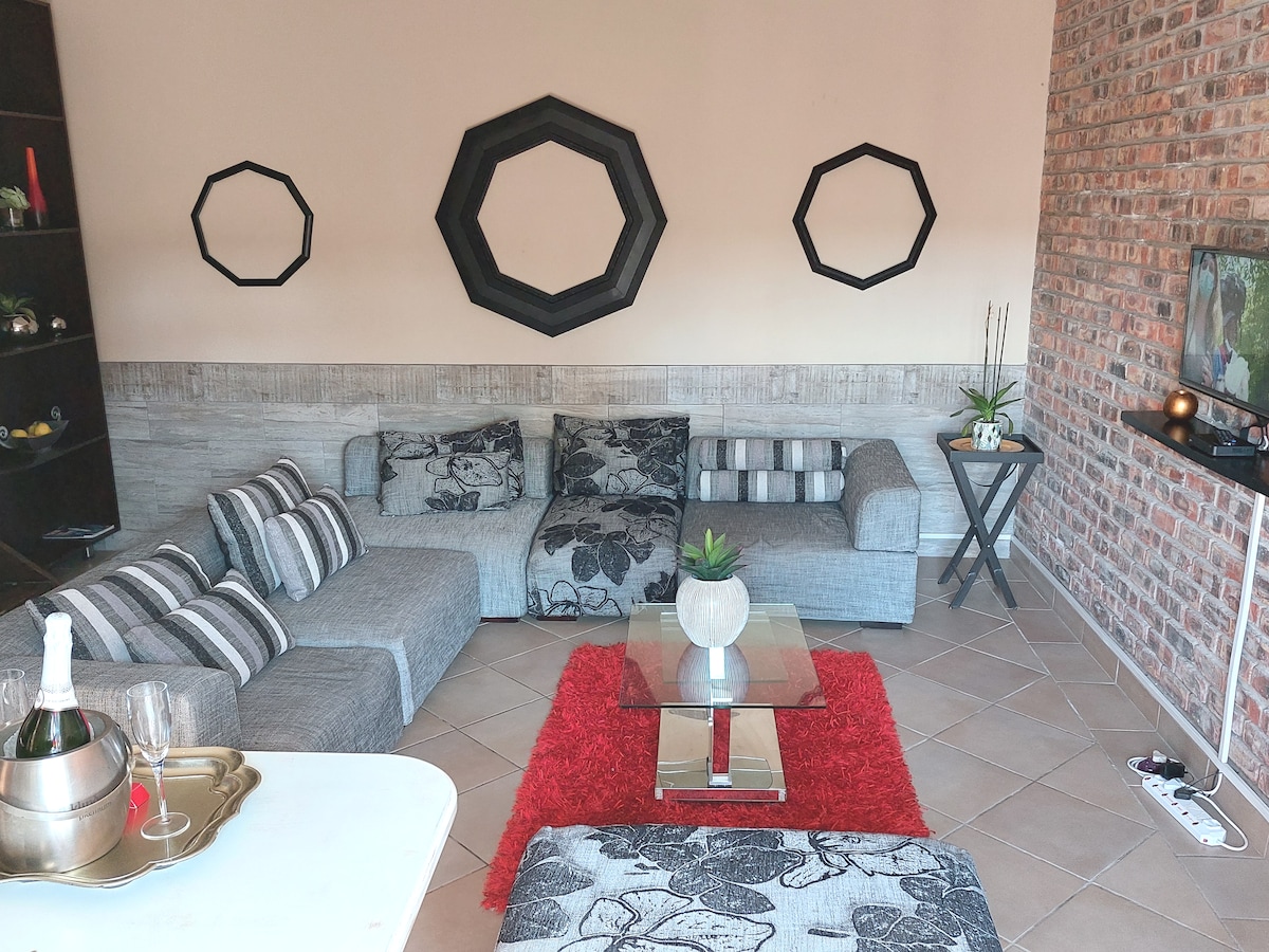 A spacious lounge area is designed with contrasting textured walls and a cozy sectional sofa adorned with patterned cushions. A glass coffee table is centered on a vibrant red area rug, complemented by decorative accessories and wall mirrors reflecting the light.