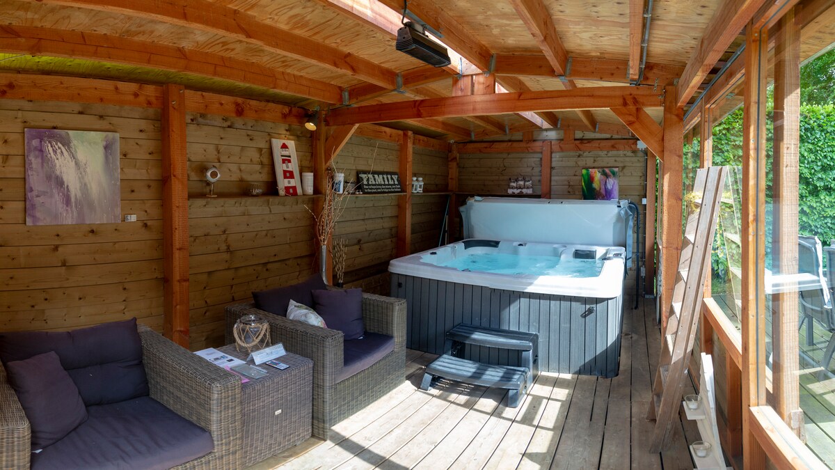 A wooden structure features a large jacuzzi surrounded by cozy seating areas. A wooden floor adds warmth, and natural light floods in through the large windows, enhancing the inviting space. Artworks adorn the walls, creating a tranquil retreat in the garden.