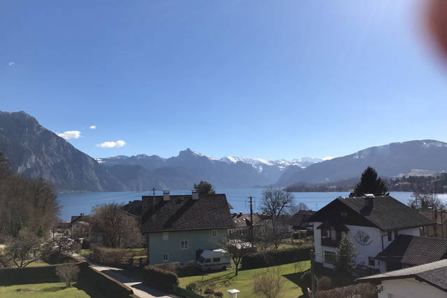 A serene landscape features a lake bordered by mountains, with snow-capped peaks in the distance. In the foreground, several houses are visible, surrounded by greenery. The clear blue sky enhances the tranquil view, creating a peaceful ambiance.