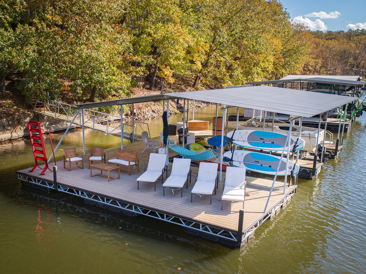 The dock features a spacious area with several lounge chairs and seating arrangements. Blue paddleboards are positioned neatly alongside, and shaded seating is available under a covered structure. Surrounding trees provide a natural backdrop, enhancing the serene water ambiance.