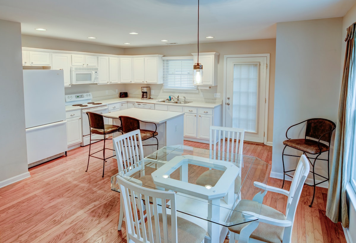 A bright and functional kitchen features white cabinetry and appliances, complemented by a central island. A glass-top dining table is surrounded by white chairs, with additional seating at a bar-height counter. Natural light enters through a nearby door leading to the back patio.