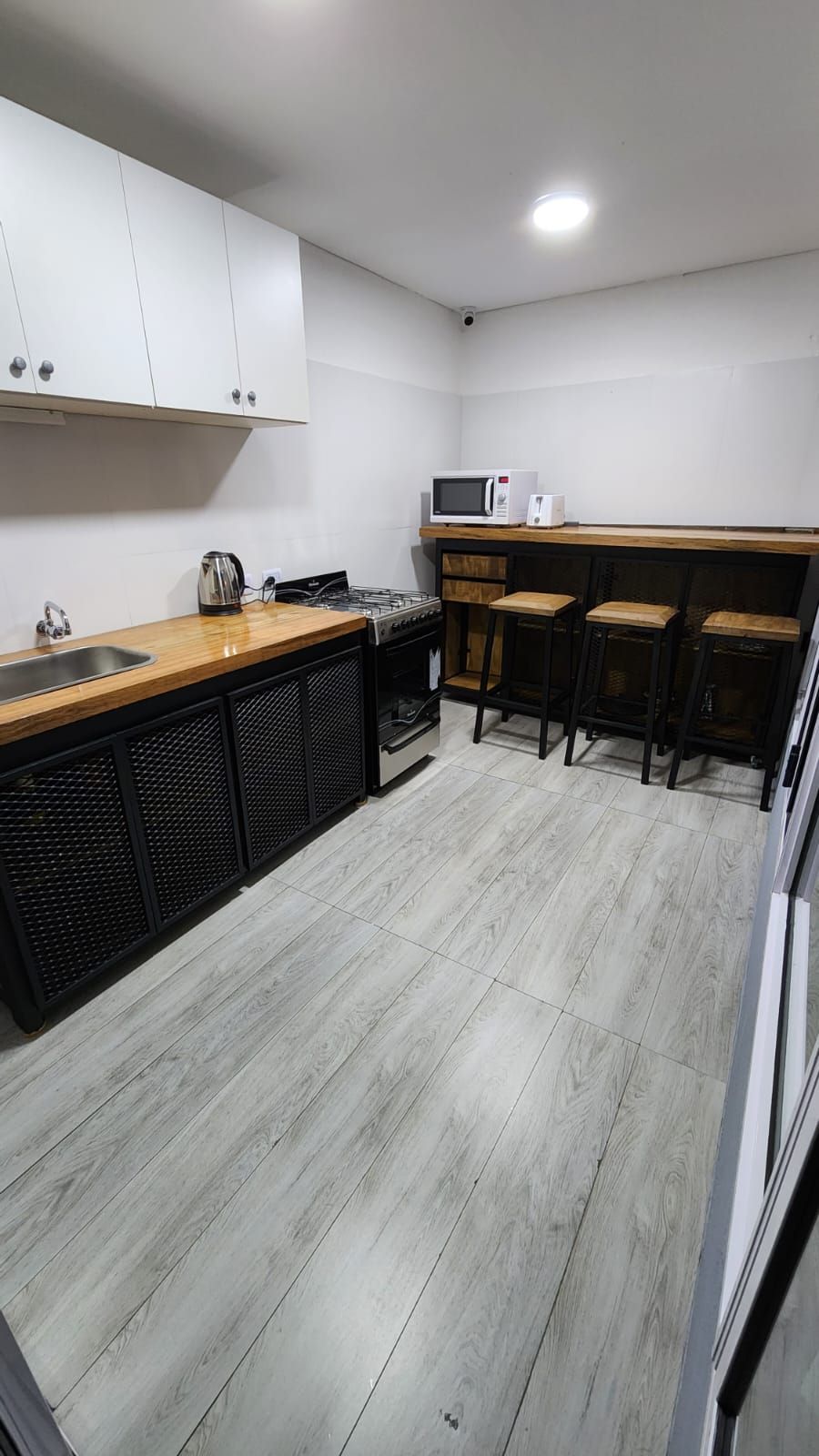 A modern kitchen area is equipped with sleek cabinetry and a wooden countertop. Essential appliances include a microwave, kettle, and electric stovetop. Four bar stools are positioned at a high counter, and light-colored flooring enhances the space's brightness.