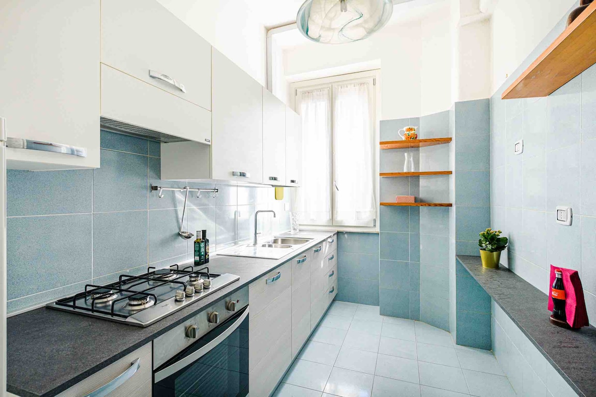 The kitchen features a modern design with light blue tile walls and a spacious countertop. A gas stove and stainless steel sink are positioned along one wall. Open shelves display a decorative object, while a small plant adds a touch of greenery to the space.