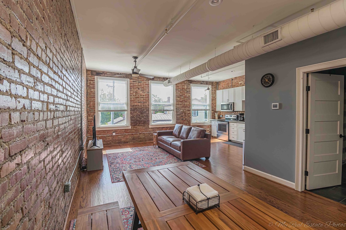An inviting living area features exposed brick walls and modern furnishings, including a spacious sofa and a wooden coffee table. Natural light enters through the large windows, enhancing the warm tones of the hardwood flooring and visible industrial accents.