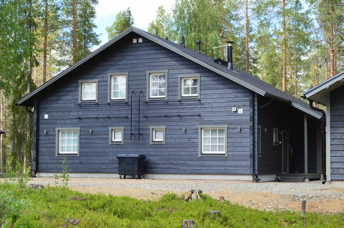 A wooden duplex apartment is shown, featuring a dark exterior with contrasting white window frames. A gravel pathway leads to the entrance, surrounded by greenery, with a trash bin placed at the front. Trees provide a natural backdrop to the property.