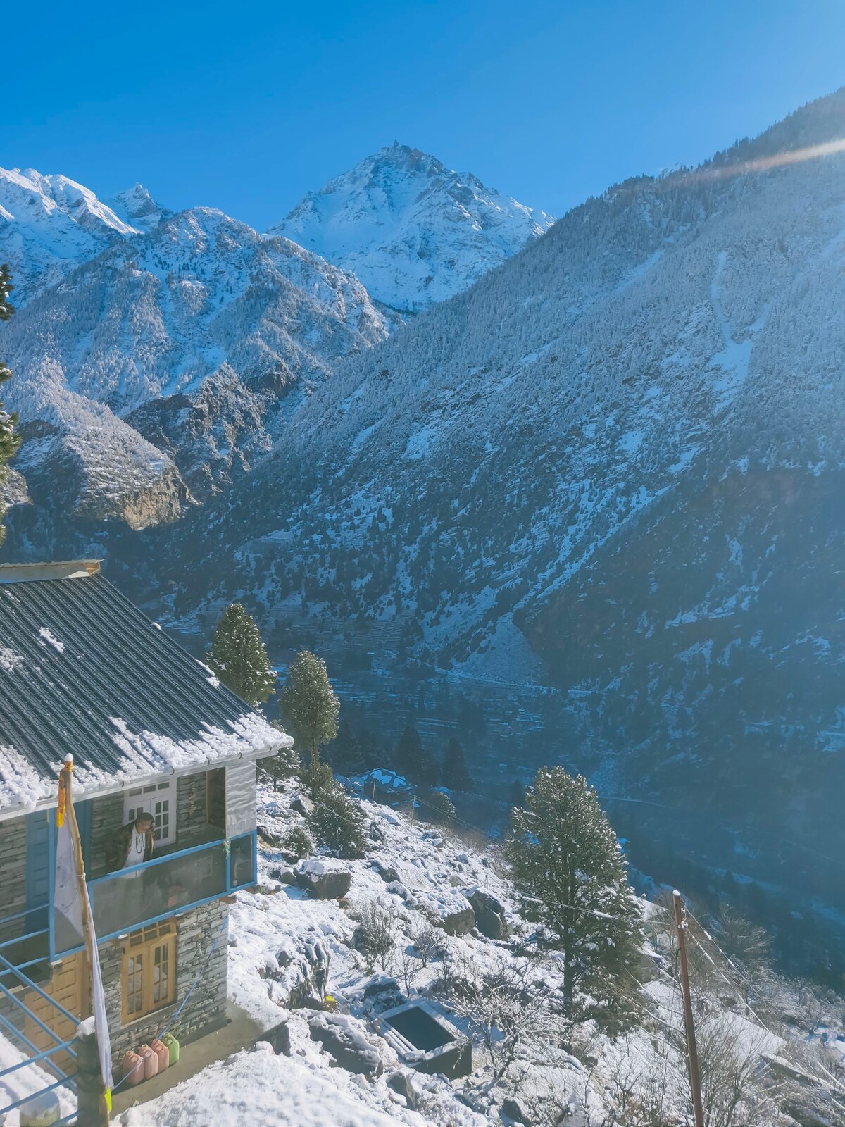 A charming eco-friendly accommodation is nestled against a backdrop of snow-covered mountains. The structure features wooden accents and large windows, allowing views of the surrounding natural landscape. Evergreen trees are visible, alongside a hint of the valley below, creating a serene atmosphere.