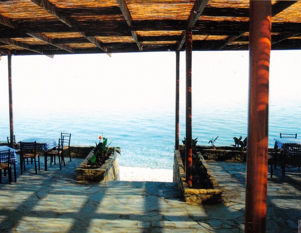A shaded outdoor dining area features wooden beams overhead and stone flooring. Tables with checkered tablecloths are positioned near the edge, alongside planters with greenery. The tranquil sea is visible beyond the seating, creating a peaceful coastal atmosphere.