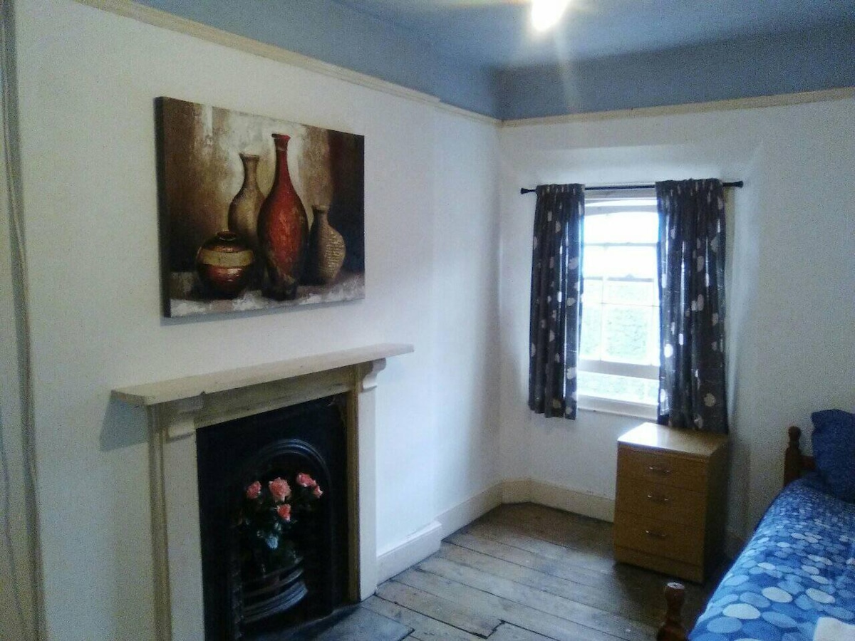 A cozy room features a decorative painting depicting vases on a light-colored wall. A wooden bed with a blue patterned bedspread is positioned adjacent to a window covered with polka-dot curtains. A small wooden chest of drawers sits beside the fireplace, which is adorned with a small floral arrangement.