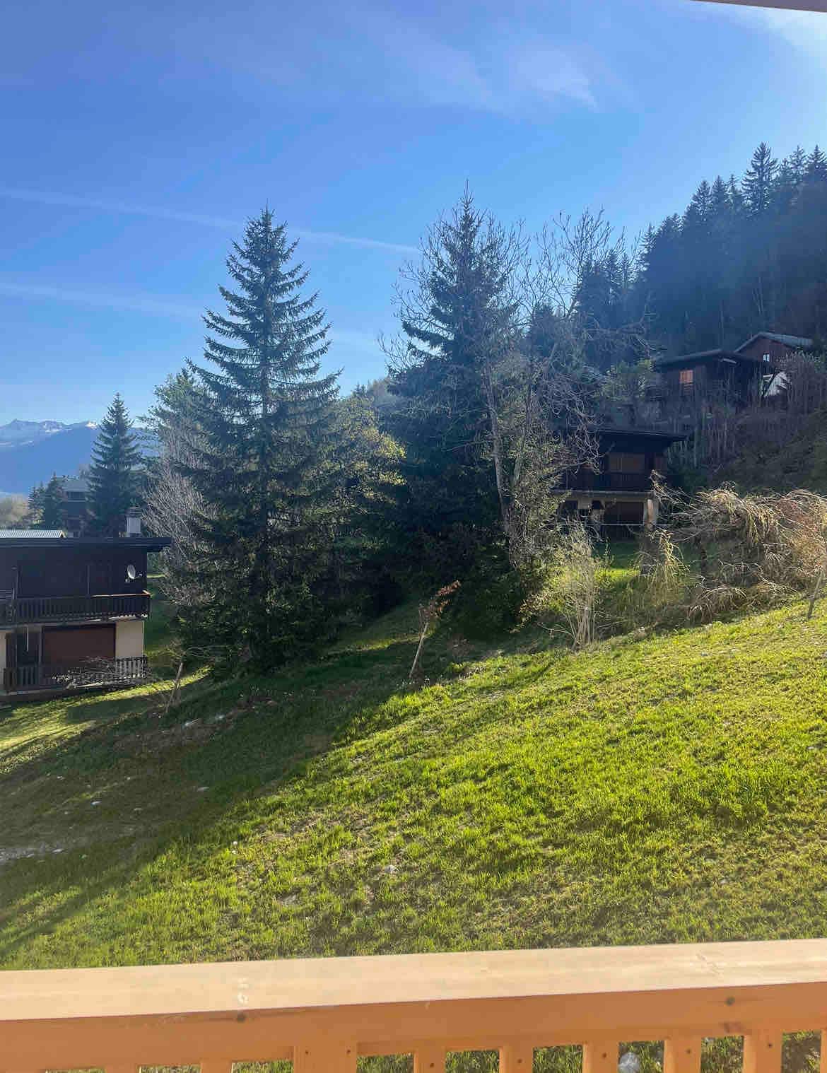 A serene landscape is visible from the balcony, featuring lush green grass and tall trees. Nearby wooden structures blend into the natural surroundings, with mountains in the distance under a clear blue sky. Soft sunlight illuminates the scene, enhancing the tranquil ambiance.