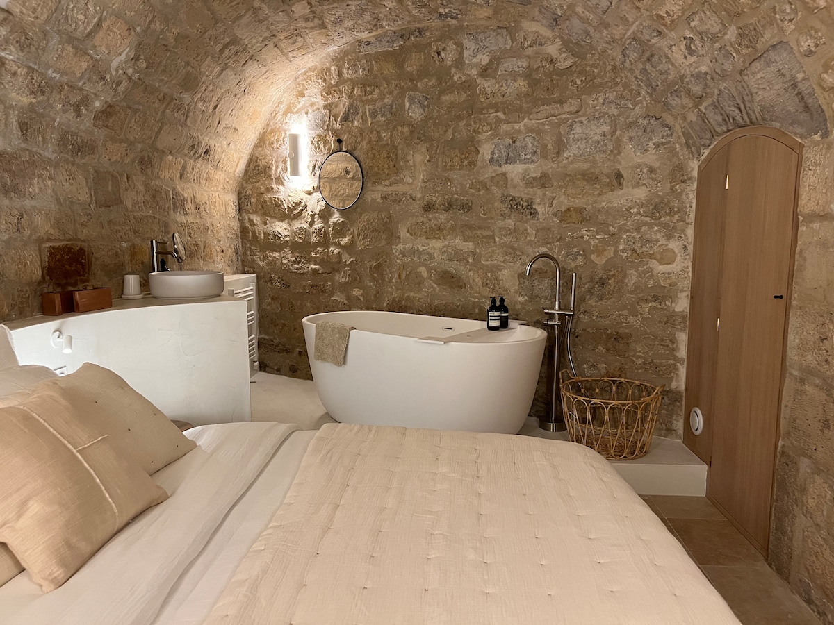 The bedroom features an arched stone ceiling and a queen-sized bed dressed in neutral linens. A freestanding bathtub is positioned for relaxation, alongside a stylish sink and mirror. Natural materials are showcased in the woven basket, enhancing the serene ambiance.