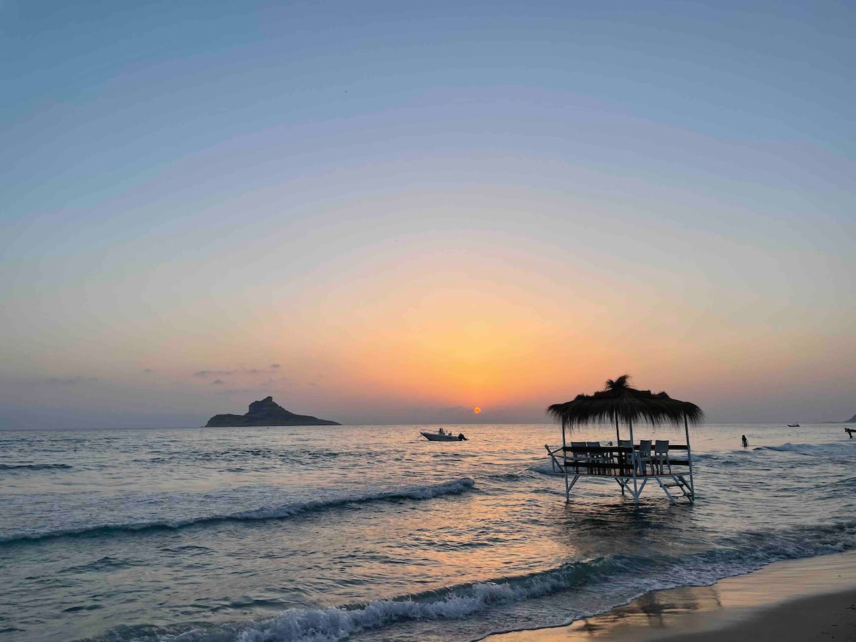 The image captures a serene coastal scene at sunset, with gentle waves lapping at the shore. A wooden structure with a thatched roof is visible over the water, with a distant rocky island silhouetted against the colorful sky. Small boats are seen on the horizon.