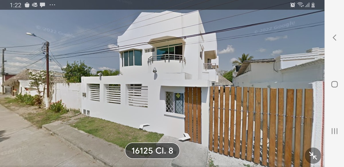 A modern two-story building is presented, featuring a clean white exterior with horizontal slats along the lower level. The upper floor includes a balcony with glass railings, and a driveway is visible alongside the property. Lush greenery is seen beyond the wooden fence.