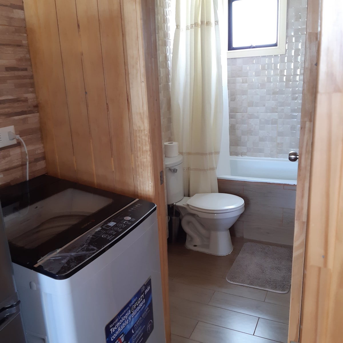 The bathroom features a washing machine beside a toilet and a bathtub with a shower. Light-colored tiles cover the walls, while a window allows natural light to enter the space. A small bath mat is positioned in front of the toilet.