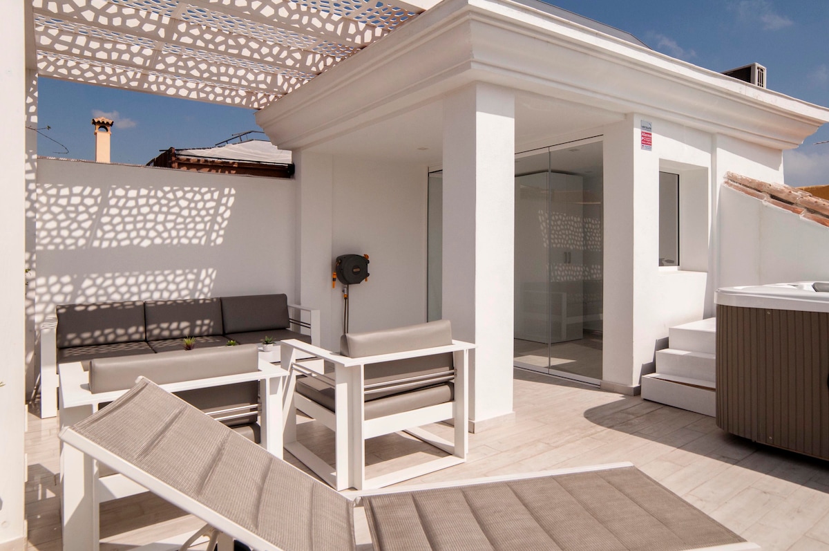 A sunlit terrace features a modern white pergola casting geometric shadows. Comfortable outdoor seating is arranged, including sleek white chairs and loungers. A private hot tub is situated in the corner, inviting relaxation. The space is designed for enjoying the outdoor sun year-round.