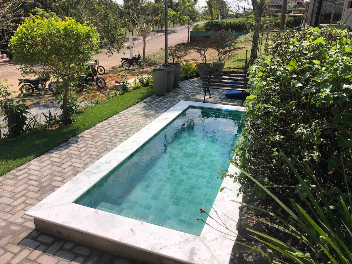 A private pool is framed by lush greenery and landscaped beds. Paved pathways lead toward the pool, while seating is provided nearby. Sunlight reflects off the clear water, creating a serene outdoor environment.