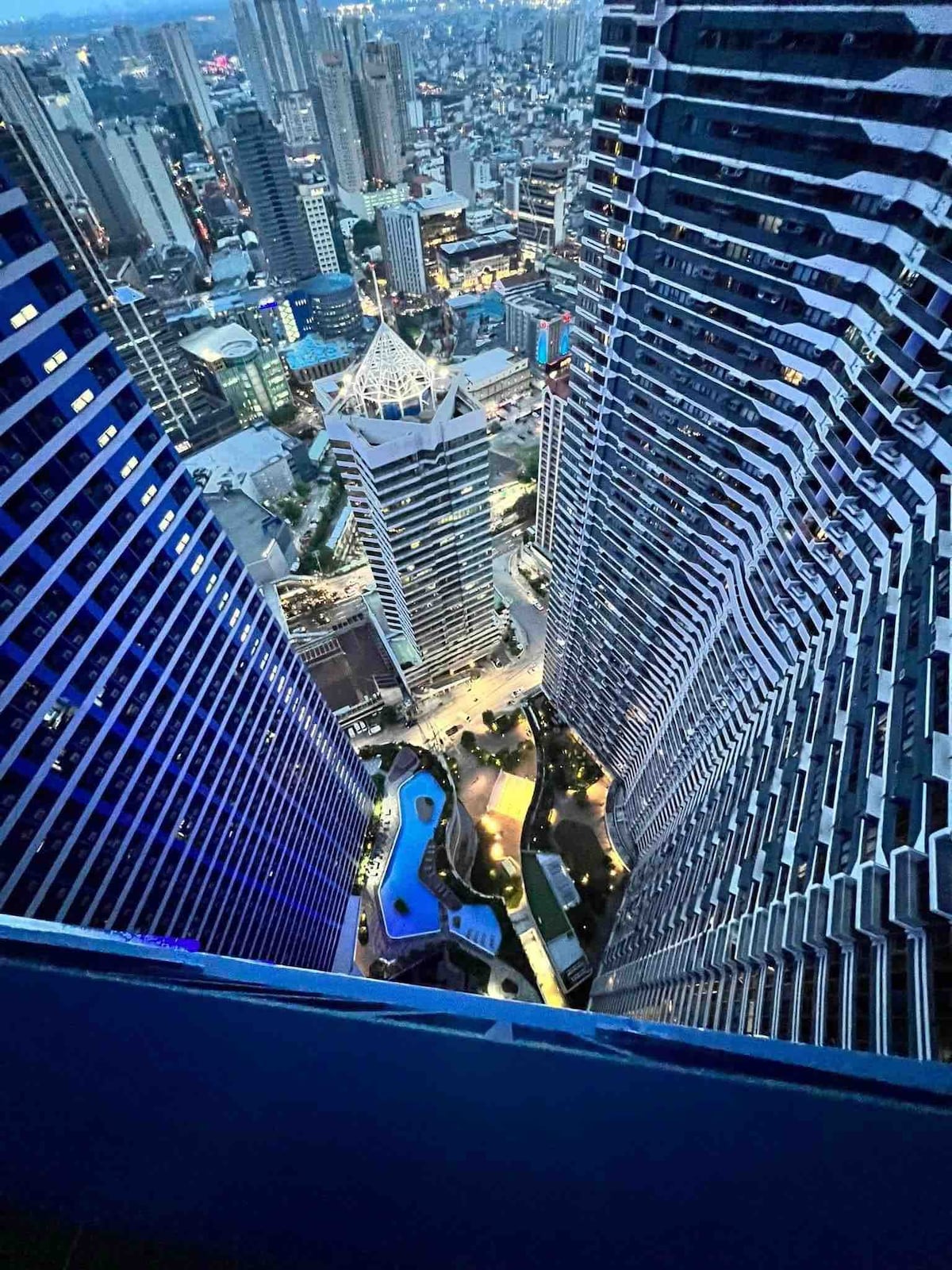 Aerial view showcases the vibrant cityscape of Makati, featuring a dynamic blend of high-rise buildings and illuminated streets. A swimming pool is visible in the lower center, surrounded by landscaped greenery, creating a serene section amid the bustling urban environment.