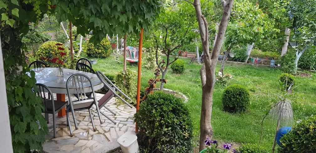 A well-maintained garden features a stone pathway surrounded by lush greenery. A round dining table with chairs is positioned under a shaded area. Trees and neatly trimmed bushes create a tranquil outdoor space, inviting relaxation and enjoyment of nature.