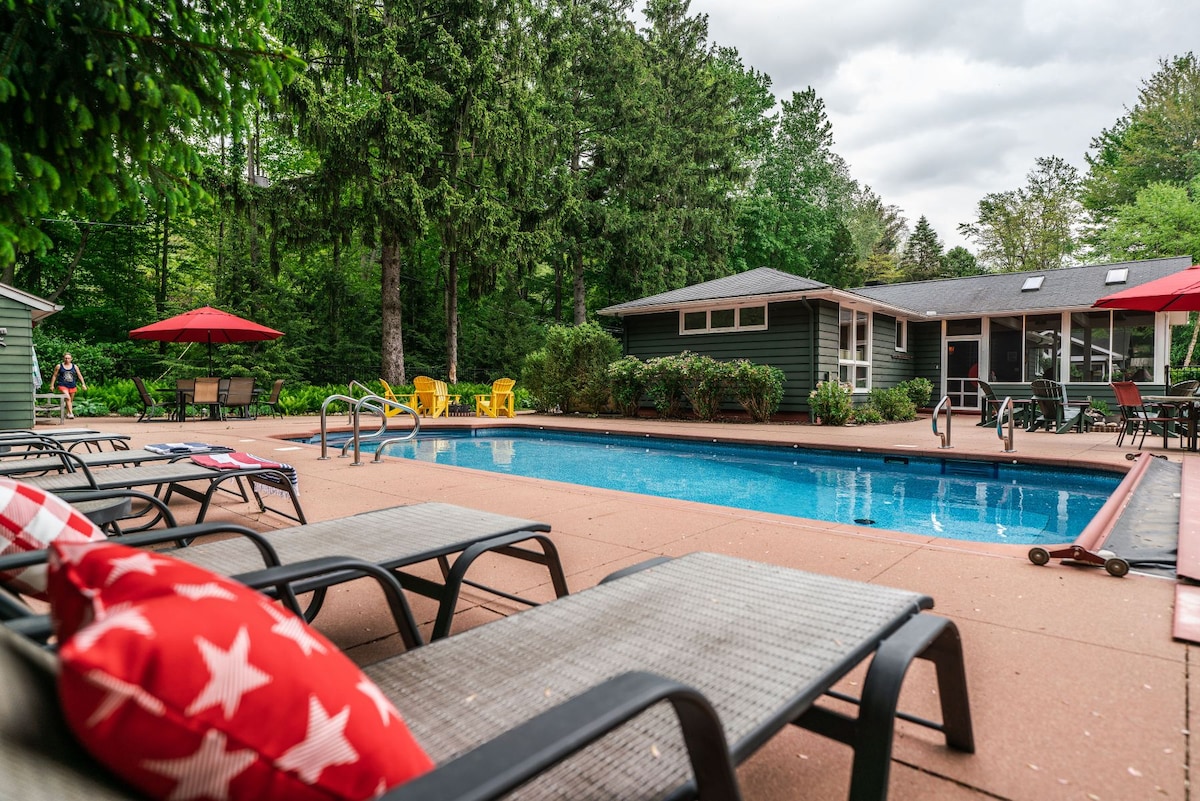 The outdoor pool area features a spacious swimming pool surrounded by lounge chairs and umbrellas. A patio is visible, along with a game room in the background, and vibrant greenery enhances the atmosphere.