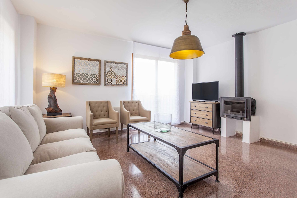 A spacious living area is presented, featuring a comfortable beige sofa and two armchairs. A wooden coffee table is centered on the polished floor. A large window invites natural light, complemented by a contemporary lamp and a wood-burning stove, creating a relaxed setting.