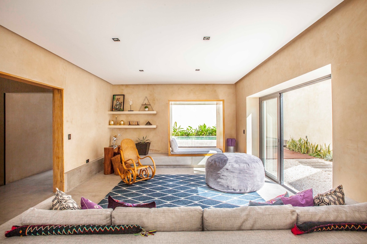 A modern living area features a light-toned, textured wall and large glass doors that provide access to an outdoor space. A comfortable beige sofa is adorned with colorful cushions. A round pouf and a rocking chair are positioned on a geometric rug, creating a relaxed atmosphere.