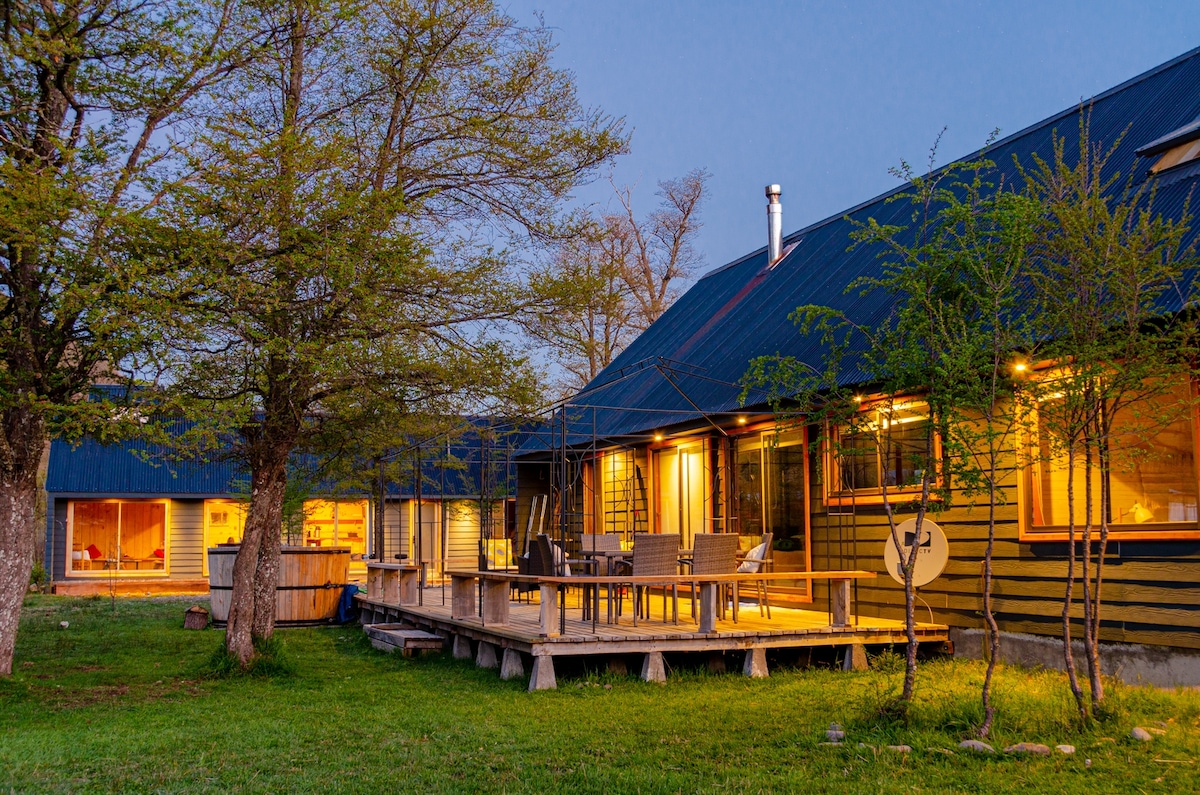 The lodge exterior is framed by trees, showcasing a spacious wooden deck with outdoor seating. Warm lights illuminate the building, creating a cozy ambiance at dusk. A hot tub is positioned nearby, inviting relaxation amidst the natural surroundings.