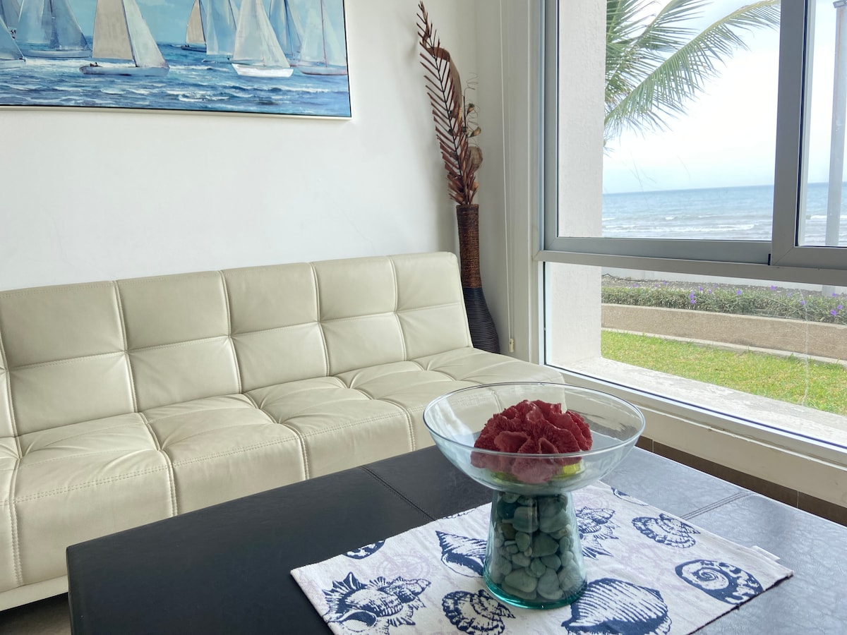 A bright living area features a light-colored sofa positioned near a large window, providing a view of the ocean. A decorative bowl filled with red fruit sits on a dark table covered with a tablecloth depicting seashells. Nearby, coastal artwork enhances the ambiance.