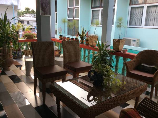 An outdoor seating area features a glass-top table surrounded by woven chairs. Potted plants add greenery to the space, which is connected to a pool visible in the background. The patio's tile flooring is arranged in a contrasting pattern of black and white.