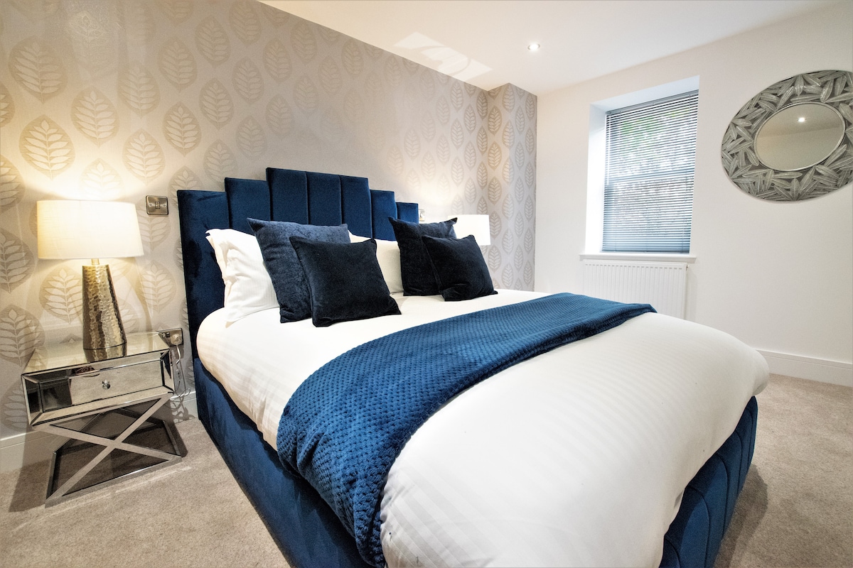 The bedroom features a spacious bed with a deep blue velvet headboard and textured bedding. Two decorative pillows enhance the bed's aesthetic. A sleek bedside lamp provides soft lighting, while a round mirror and a window with sheer blinds are visible, complementing the room's design.