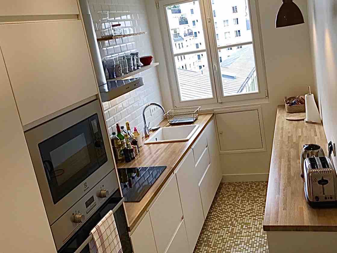 A modern kitchen is featured with white cabinets and a wooden countertop. A sink is positioned under a window, providing natural light. Cooking appliances, including an oven and stove, are visible, along with a few spices and kitchen utensils organized on shelves.