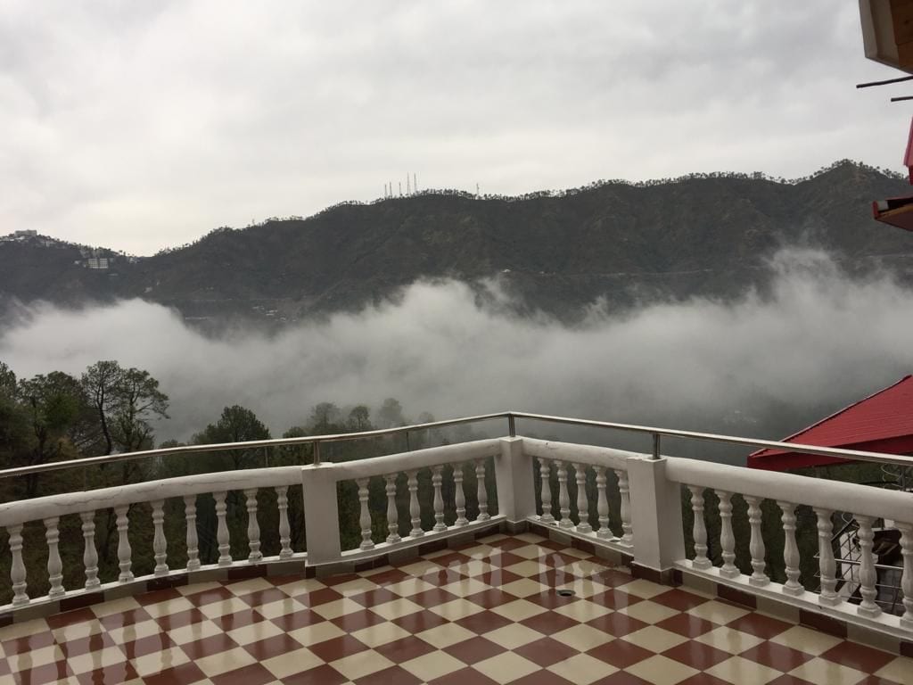 An open balcony features a patterned tile floor and a low stone railing, providing a sweeping view of mist-covered mountains. The landscape, with dense greenery, creates a serene atmosphere under overcast skies.