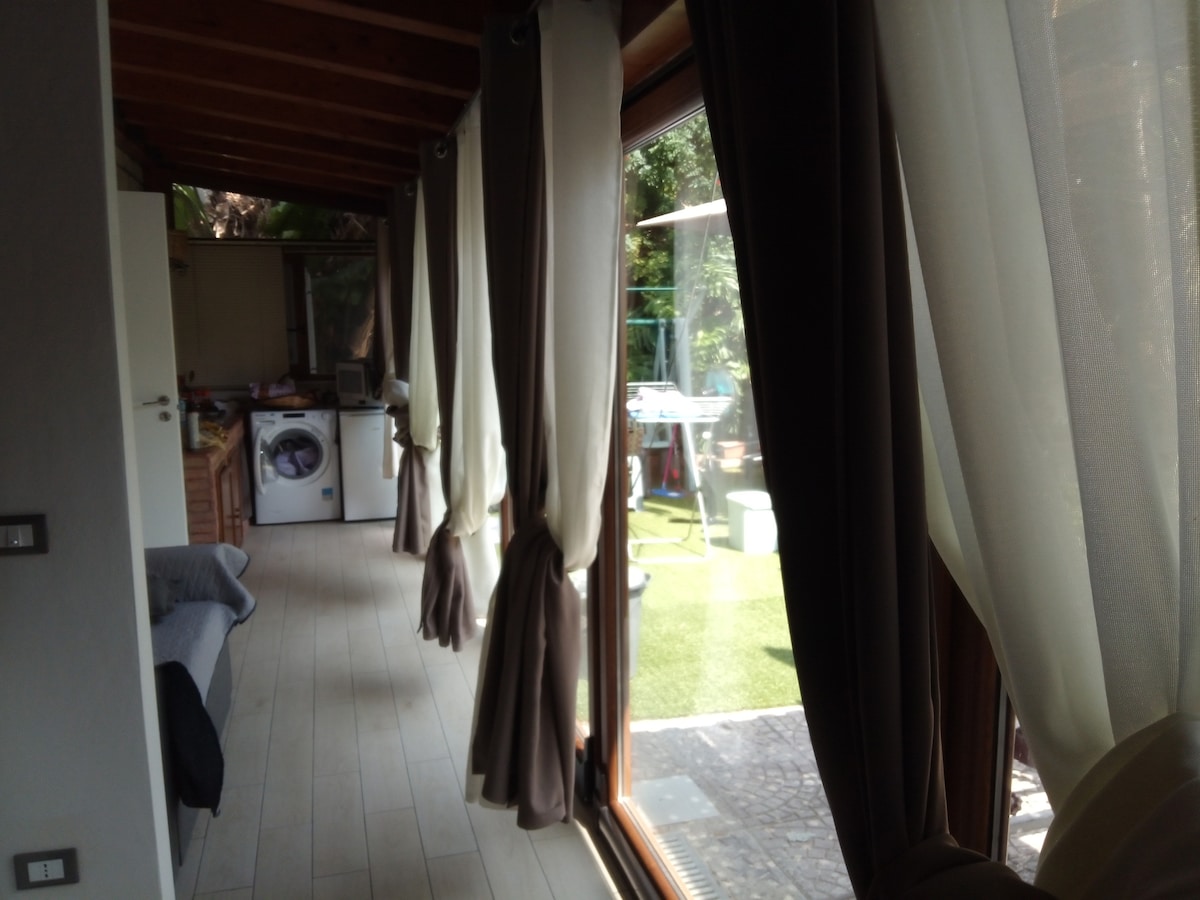 Natural light fills the spacious interior, accentuated by large glass doors that lead to the private exterior area. Elegant curtains frame the view, while a laundry area is partially visible in the background. The floor is lined with light-colored planks, contributing to a welcoming ambiance.
