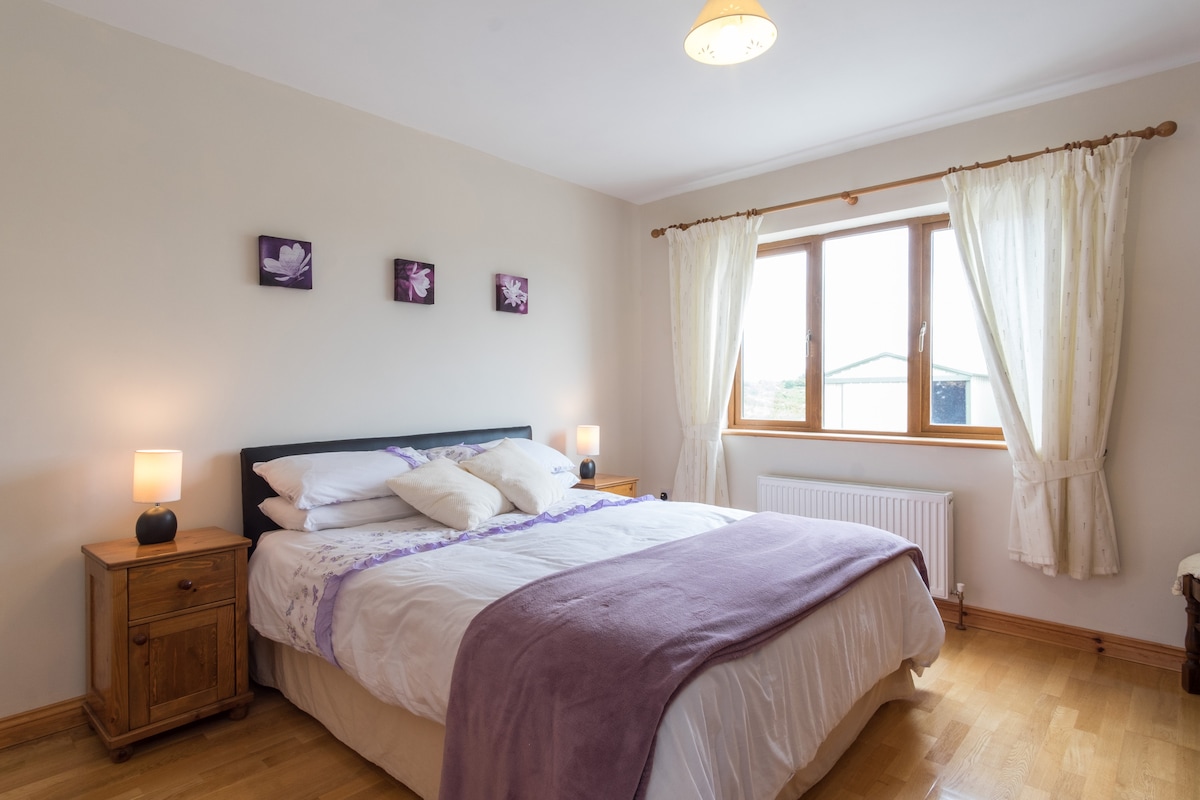 A well-lit double room features a spacious bed with white linens and a lavender throw. Bedside tables with lamps are positioned on either side. A large window with sheer curtains provides natural light and a view of the outdoors. Wall art complements the calming color scheme.