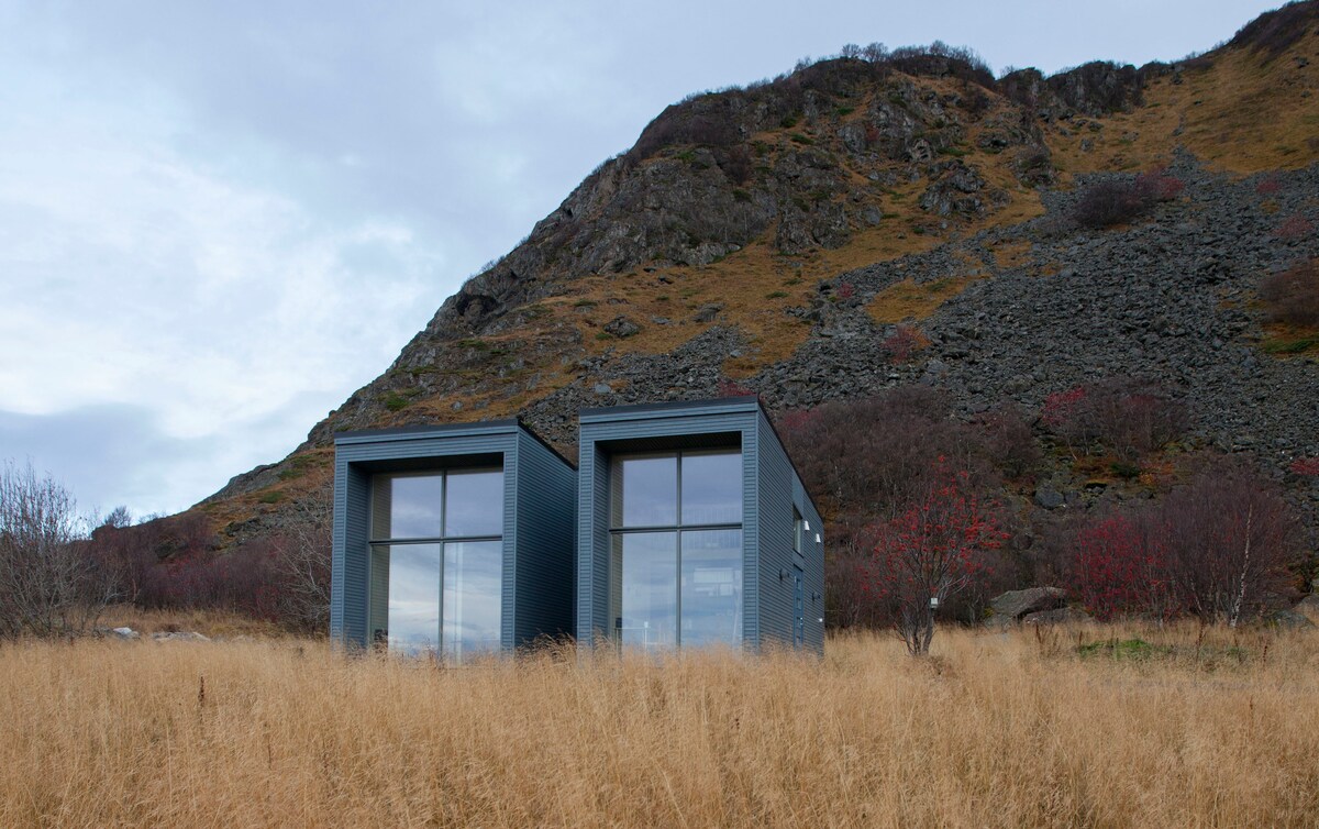 The modern structure features two adjoining units with large glass windows, set against a backdrop of rocky terrain and gently sloping hills. Tall grass sways in the foreground, adding a natural element to the scene.
