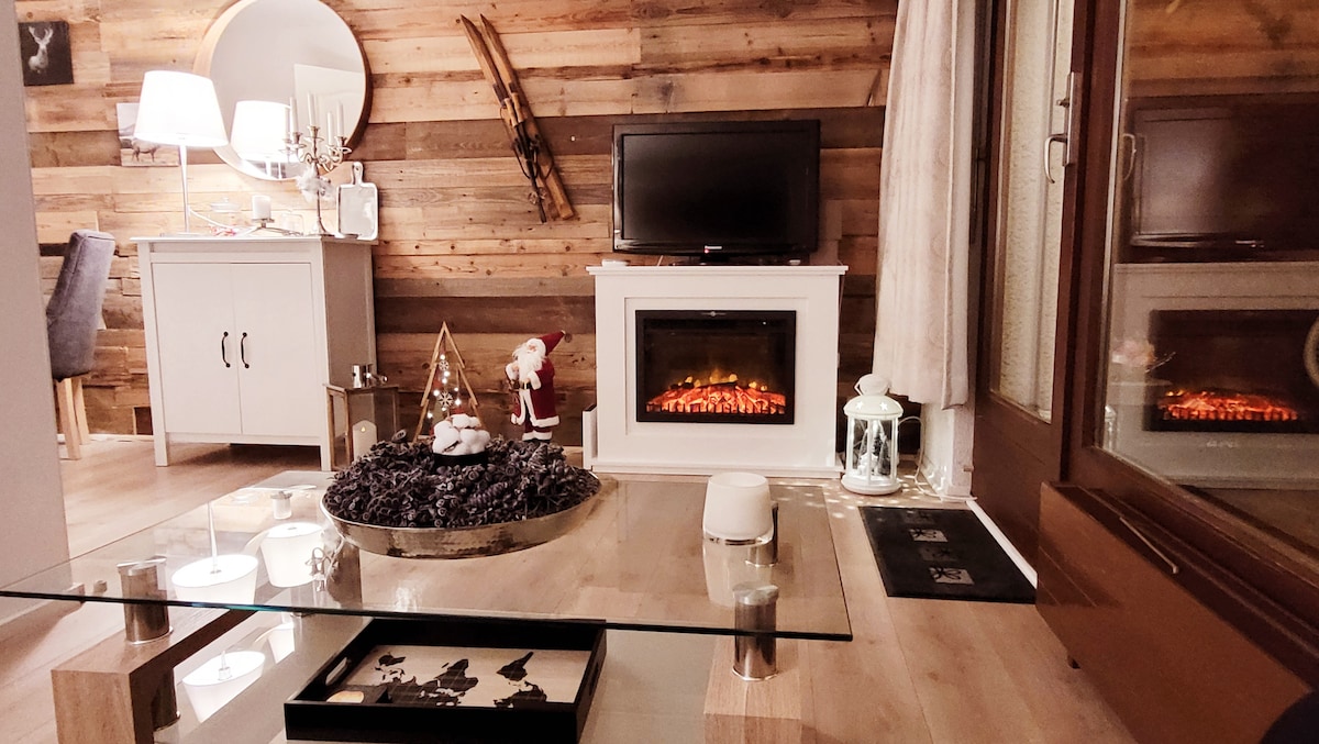 A cozy living area features a low glass table surrounded by wooden accents. A television sits atop a modern fireplace, with a decorative arrangement including candles and a small Christmas tree. Natural light filters through a nearby window, enhancing the inviting atmosphere.