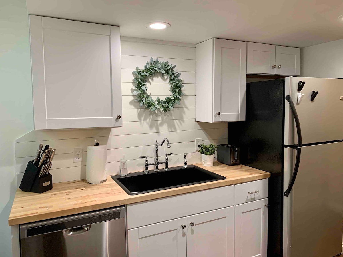 A modern kitchen features a sleek black sink set against a wooden countertop. Upper cabinets provide storage, while a green wreath adds a touch of decor above the sink. A stainless steel refrigerator and a dark refrigerator are positioned side by side for convenience.