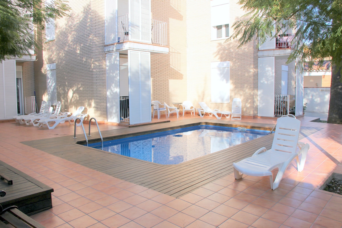 A shared swimming pool is framed by a spacious tiled area, with sunlight reflecting off the water. Lounge chairs are positioned around the pool, offering space to relax. Surrounding tall trees provide shade, enhancing the tranquil environment of the pool area.
