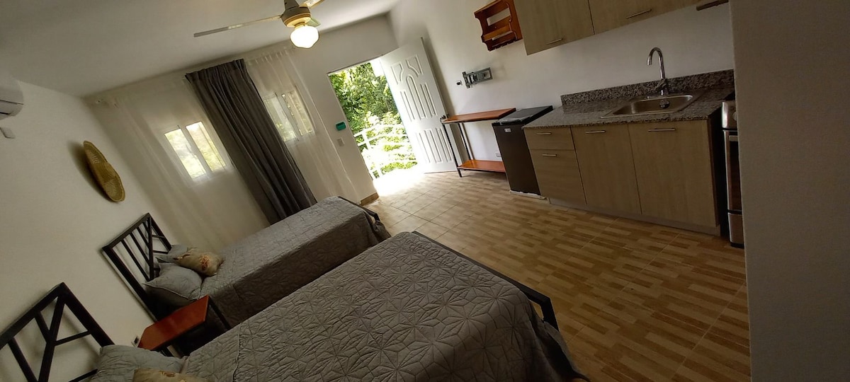 A well-lit room features two spacious beds with simple bedding. A small kitchenette, including a sink and refrigerator, is situated against one wall. Natural light enters through glass doors leading to an outdoor space, complemented by soft curtains and a ceiling fan.