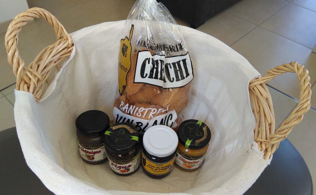 A woven basket holds a selection of local products, including jars of spreads and a package of traditional bread. The bread is prominently labeled, and various flavored jars are neatly arranged, each featuring colored tags that indicate their contents, all set against a neutral background.