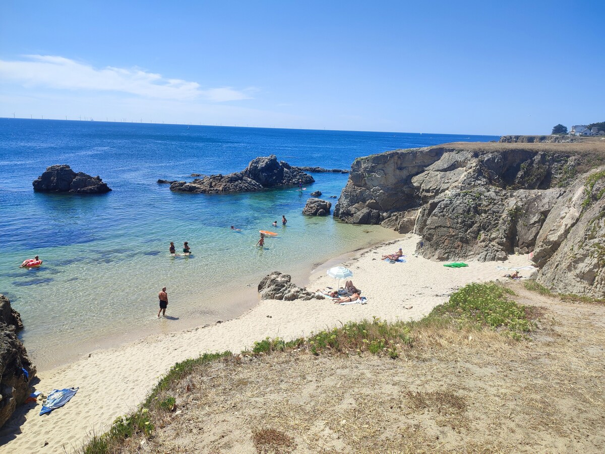 A secluded sandy beach is framed by rocky outcrops and calm blue waters. Sunbathers are seen enjoying the sun, while a few individuals swim nearby. The clear sky above and gentle waves create a peaceful seaside atmosphere.
