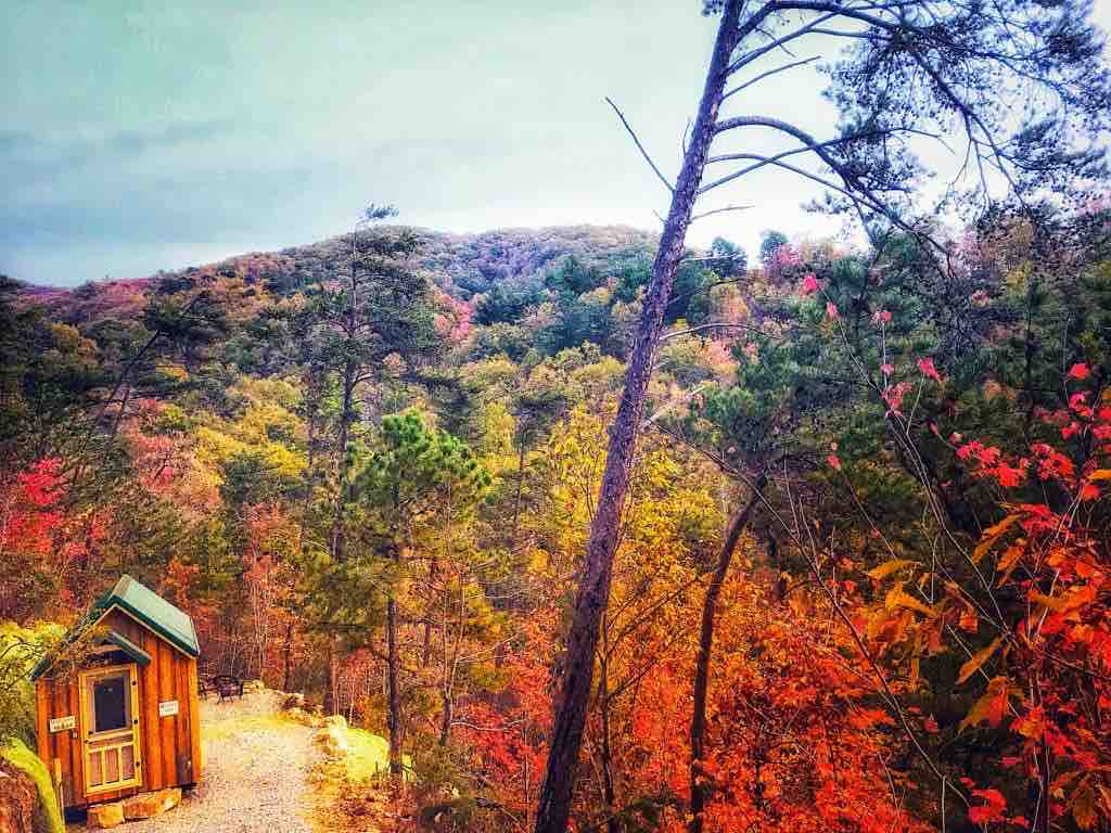 A tiny cabin is nestled along a gravel path surrounded by vibrant autumn foliage. The landscape features a tapestry of red, orange, and green leaves, set against rolling hills under a cloudy sky.