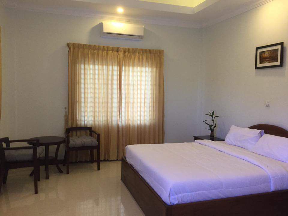 A spacious room features a large bed with crisp white linens and a minimalist wooden frame. Natural light filters through sheer curtains covering two windows. A small seating area includes a wooden table and two chairs, providing a comfortable space for relaxation.