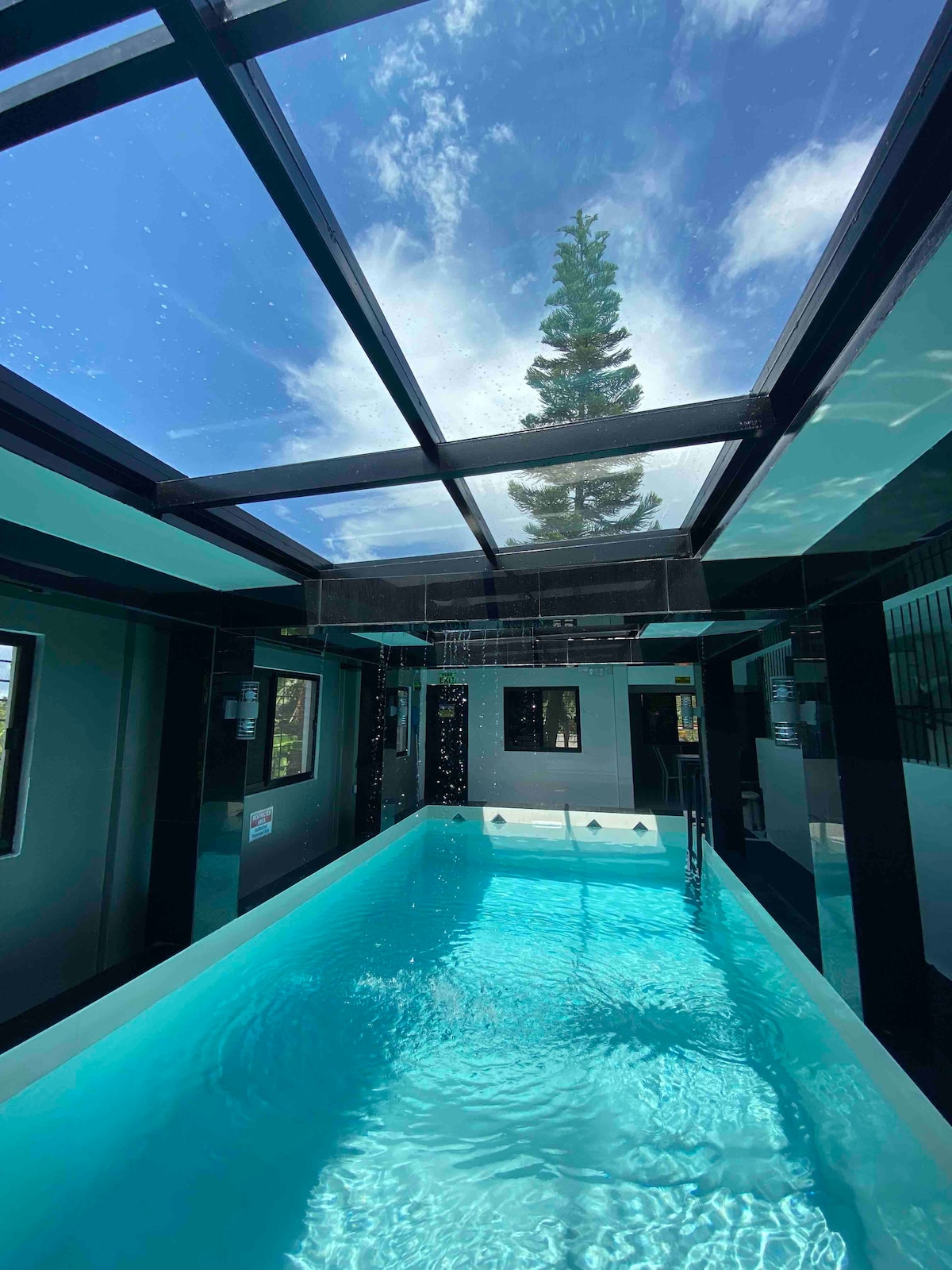 An indoor pool is showcased beneath a glass ceiling, offering a view of the sky and surrounding greenery. The clear water's blue hue reflects the natural light, creating a serene environment. Black and white accents enhance the modern design of the space.
