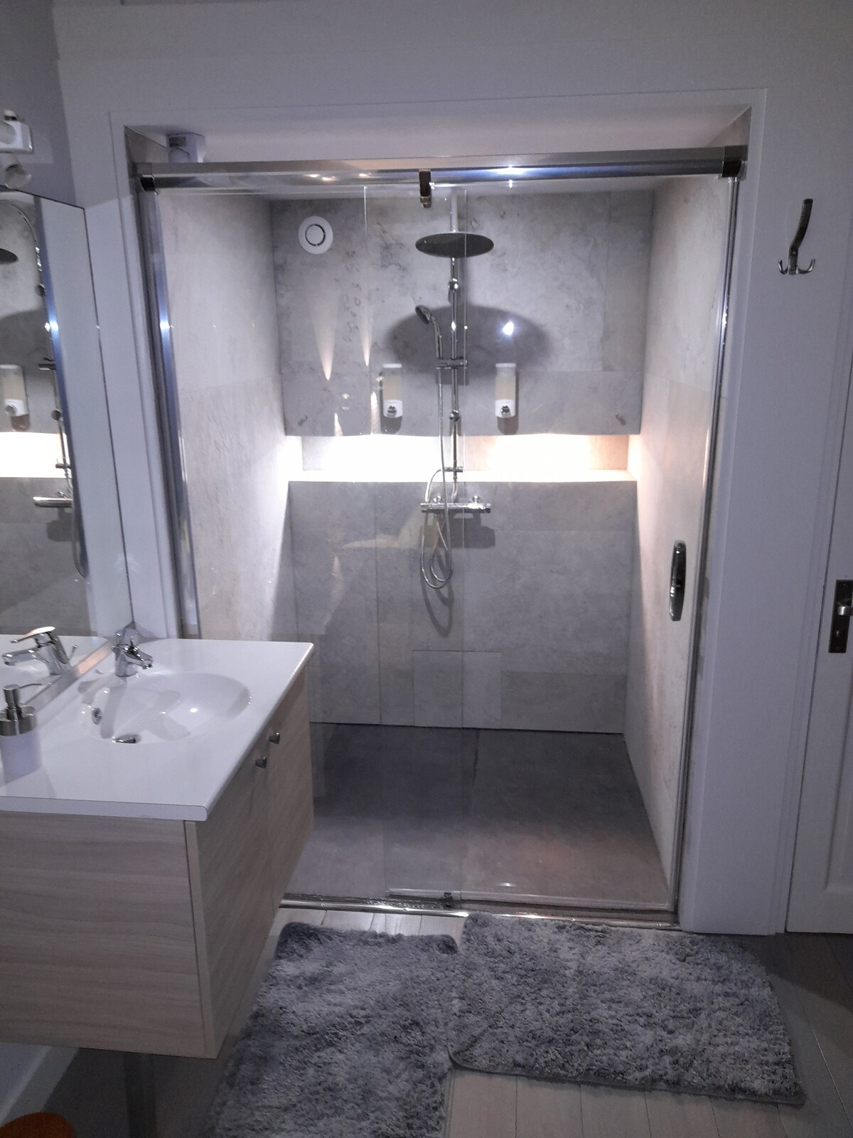 A modern bathroom features a glass-enclosed shower with sleek, light-colored tiles. A rainfall showerhead is mounted above, while built-in lighting highlights the space. A simple vanity area with a white sink and storage cabinet is positioned nearby, complemented by soft bath mats on the floor.