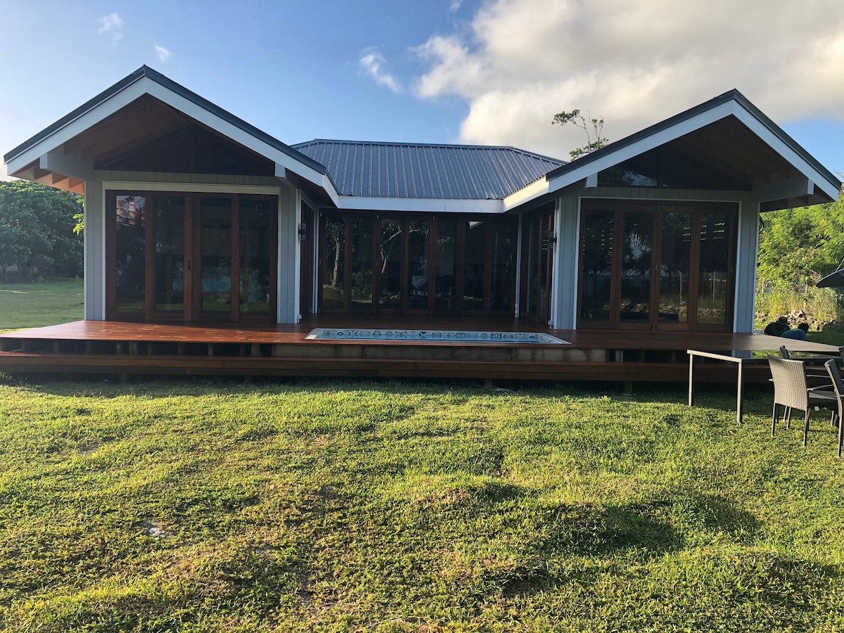 A modern villa is highlighted by a spacious deck and large glass doors that invite natural light. The structure features a sloped roof, and surrounding green grass enhances the outdoor setting, providing a sense of openness and tranquility.