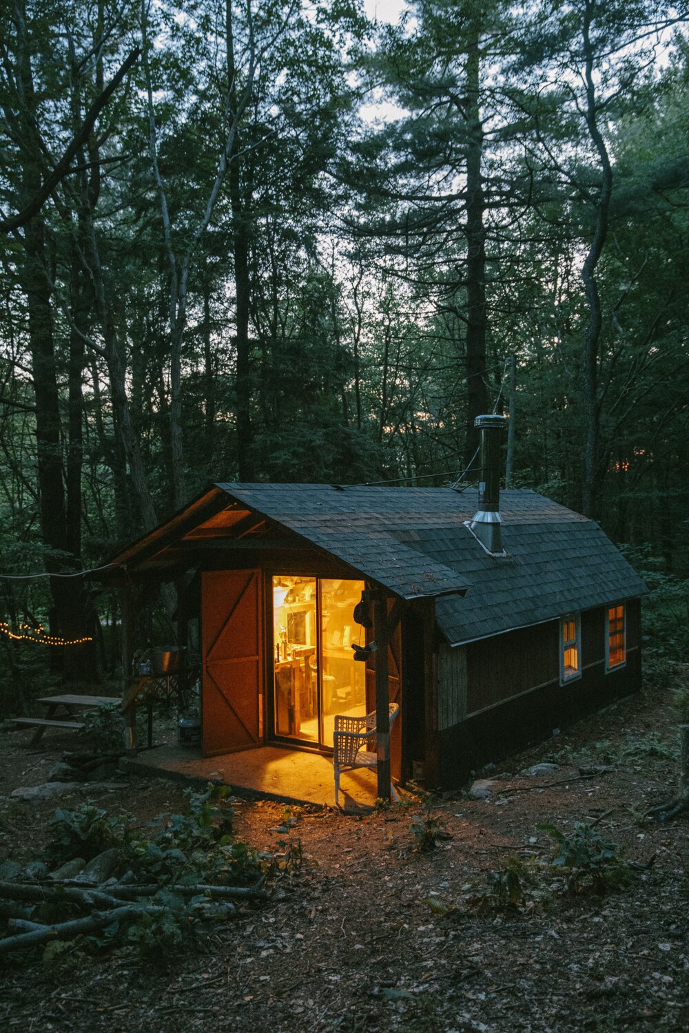 A cozy cabin nestled among towering trees in a wooded setting. Warm light spills from the windows, illuminating the entrance. The rustic exterior features a sloped roof and a chimney, surrounded by a natural landscape of forest floor and greenery.