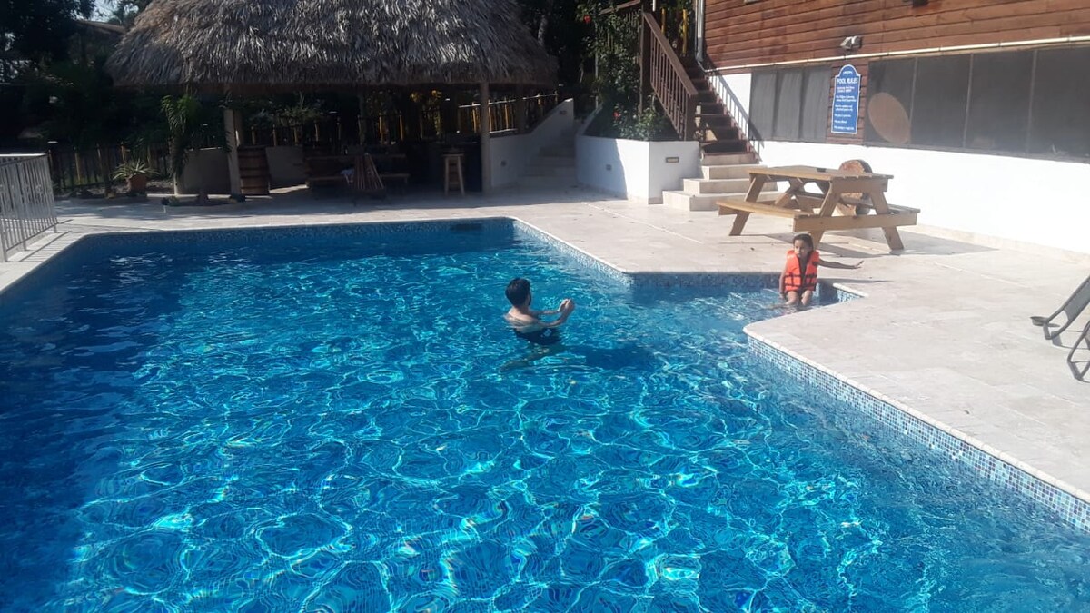 A spacious swimming pool is surrounded by a well-maintained area, featuring clear blue water. A shaded cabana stands nearby, and a picnic table is positioned on the pool deck. Sunlight creates a shimmering effect on the water's surface, providing a relaxing ambiance.