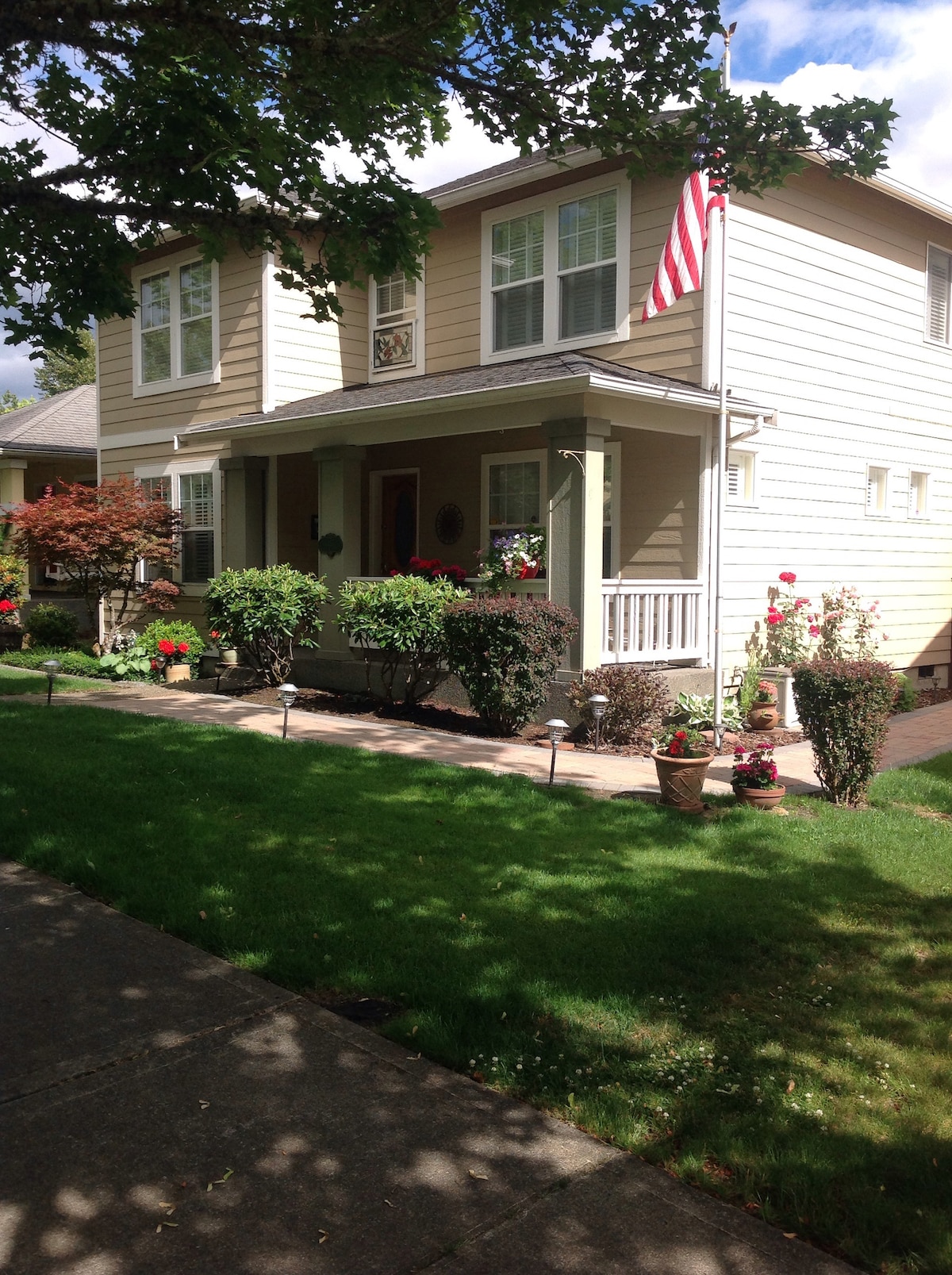 The exterior of a two-story home is framed by lush, green lawns and well-maintained flower beds. Several potted plants add color to the front porch, and an American flag gently waves above the entrance, inviting guests to explore further.